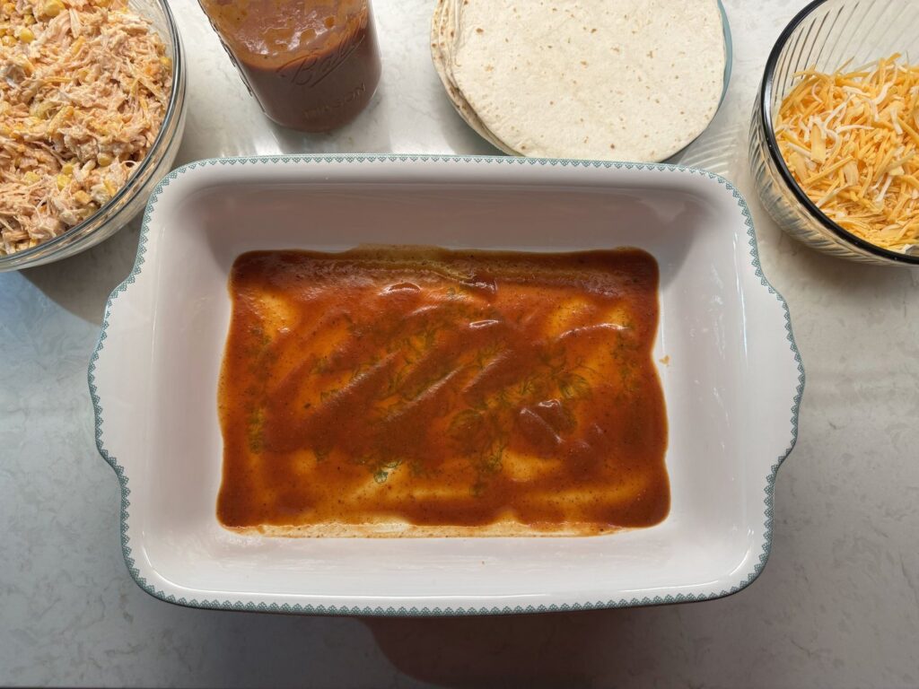 A thin layer of red enchilada sauce spread across the bottom of a white baking dish