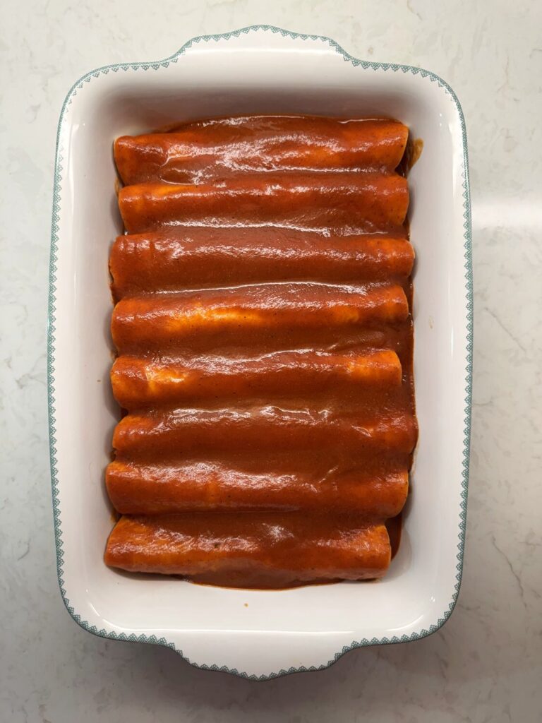 Rolled enchiladas completely covered in red enchilada sauce in a white baking dish