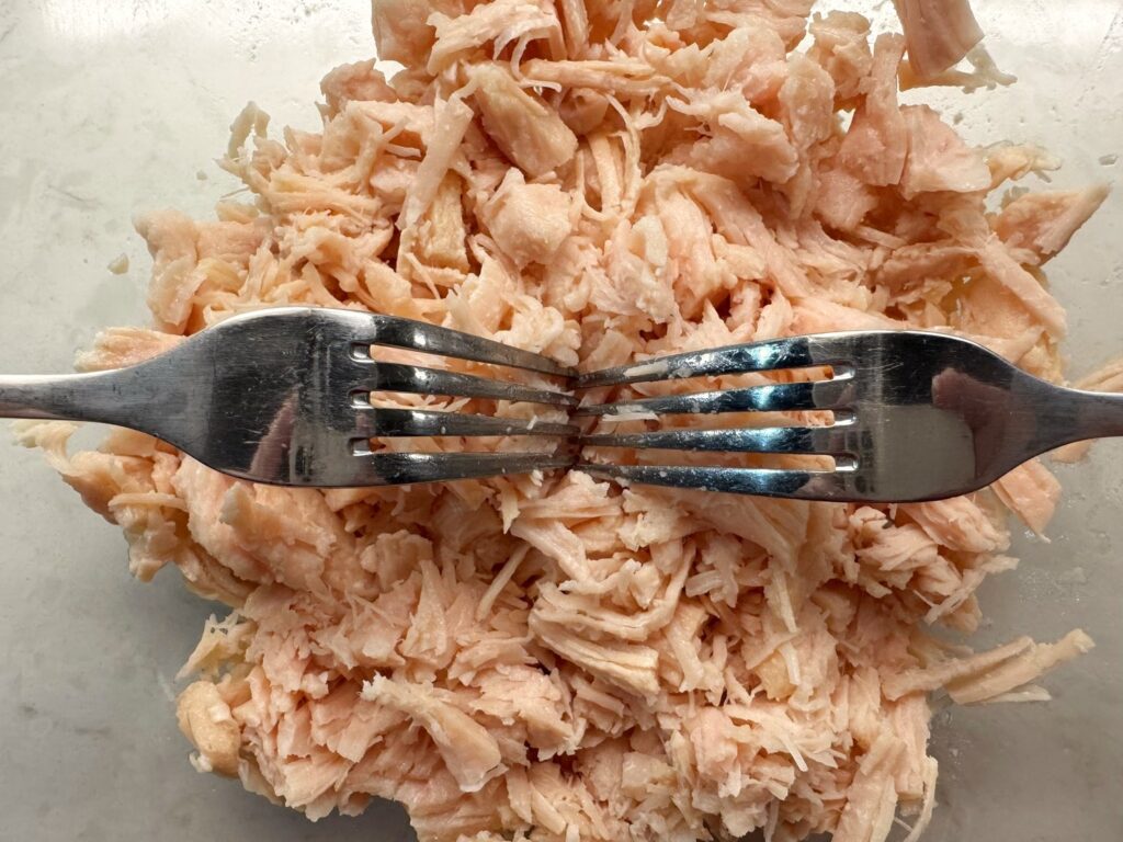 Two forks shredding drained canned chicken in a glass mixing bowl