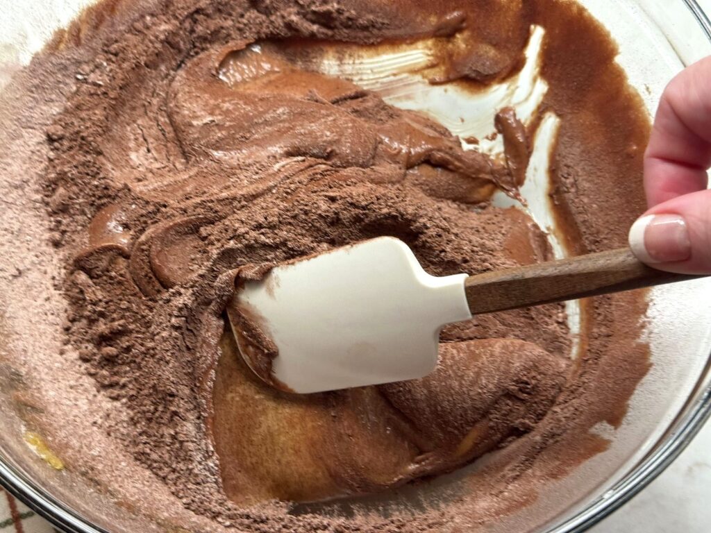 A white spatula folding flour and cocoa into chocolate brownie batter in a glass bowl