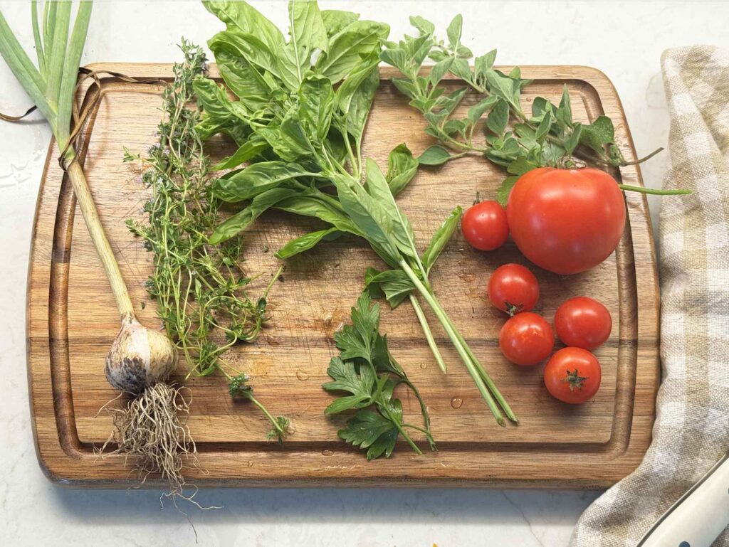 A cutting board with fresh-cut vegetables: garlic, basil, oregano, parsley, thyme, and tomatoes