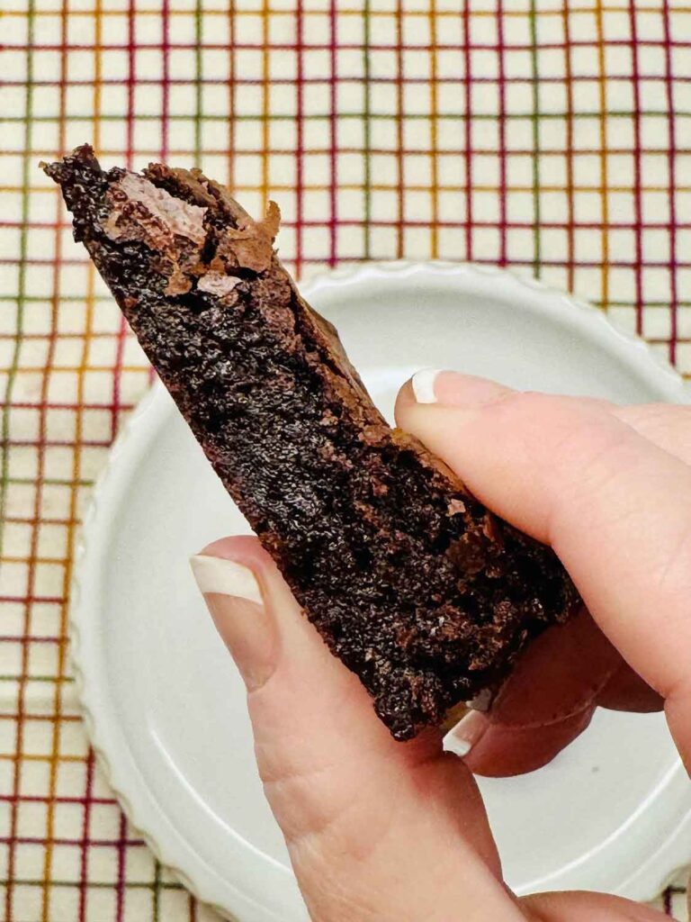 A hand holding a brownie broken in half, showing the dense, fudgy inside