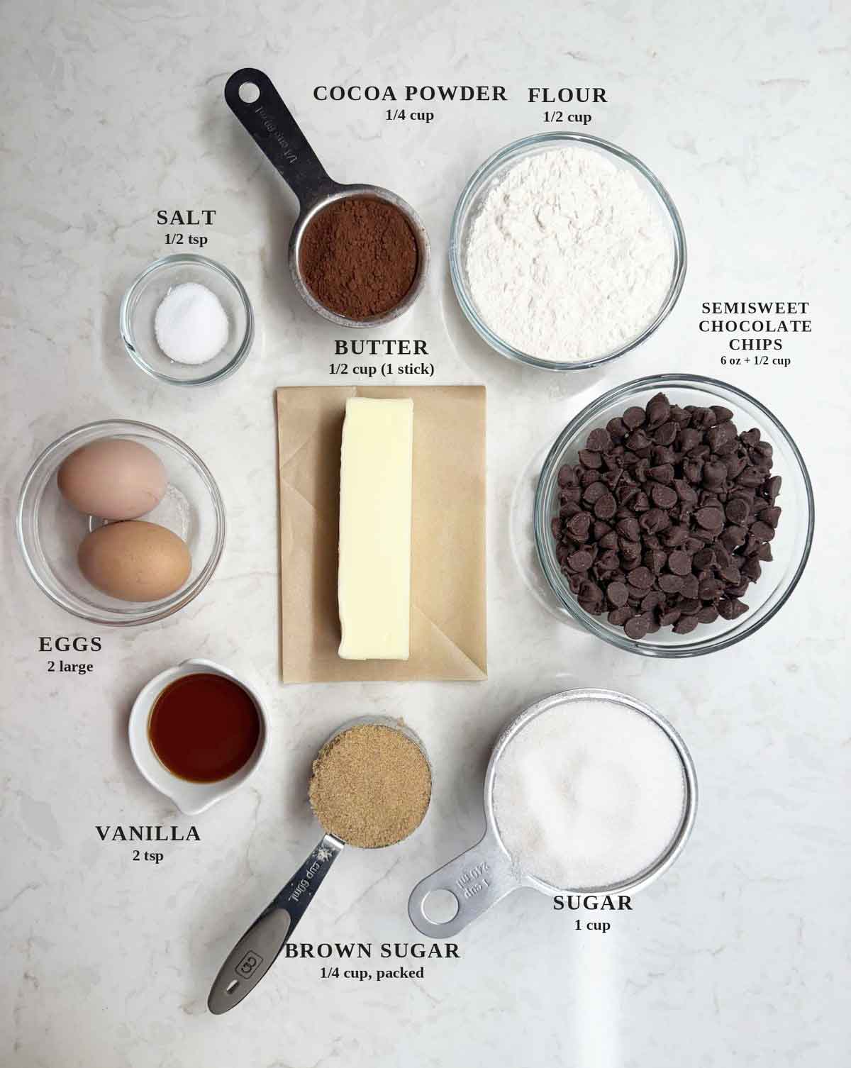 Labeled ingredients for fudgy brownies: cocoa powder, flour, salt, butter, semisweet chocolate chips, eggs, vanilla, brown sugar, and granulated sugar on a white counter