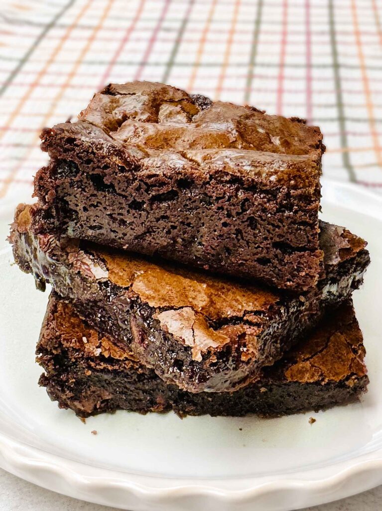 A stack of three fudgy homemade brownies with shiny crackly tops on a white plate