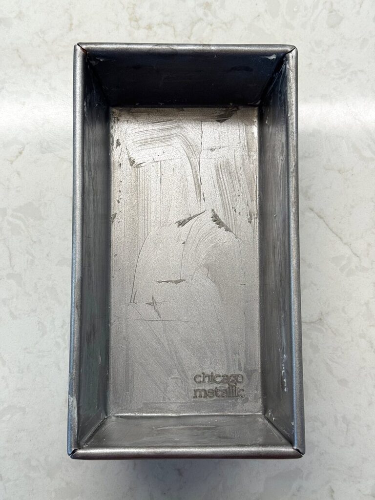 A greased 9 x 5-inch metal loaf pan with visible streaks of butter on the bottom and sides