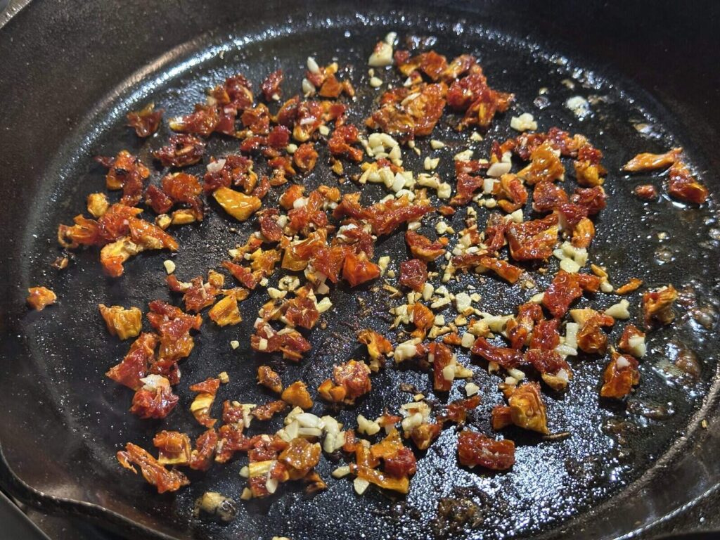 Chopped sun-dried tomatoes and minced garlic cooking in a cast iron skillet
