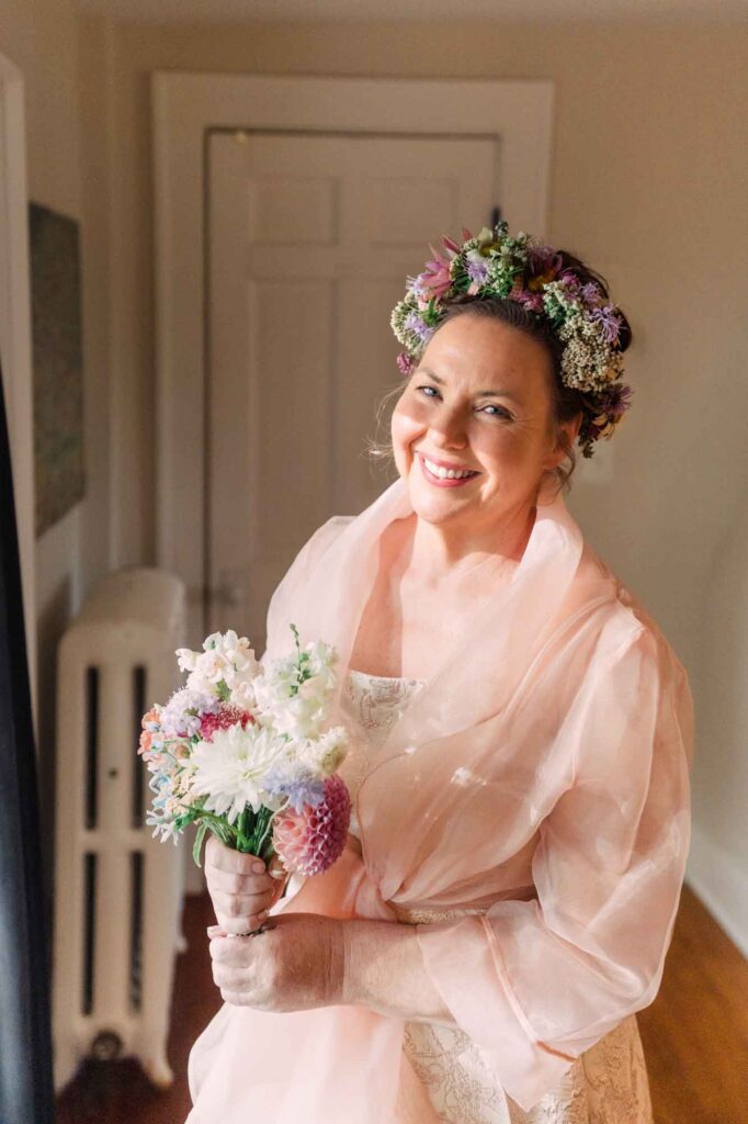 Jennifer Maker in a pink dress holding her wedding bouquet and wearing a crown of wildflowers from her cottage garden