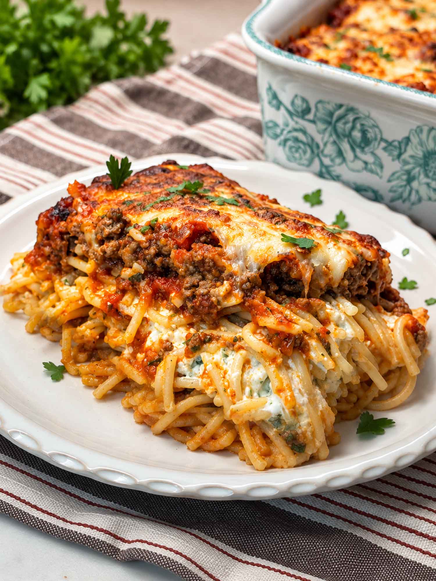 A serving of million dollar spaghetti on a white plate with fresh parsley and the baking dish behind it.