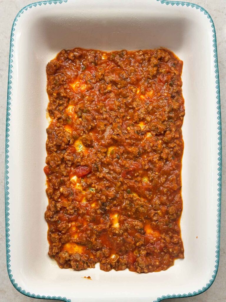 A thin layer of ground beef spaghetti sauce spread across the bottom of a white 9x13 baking dish.