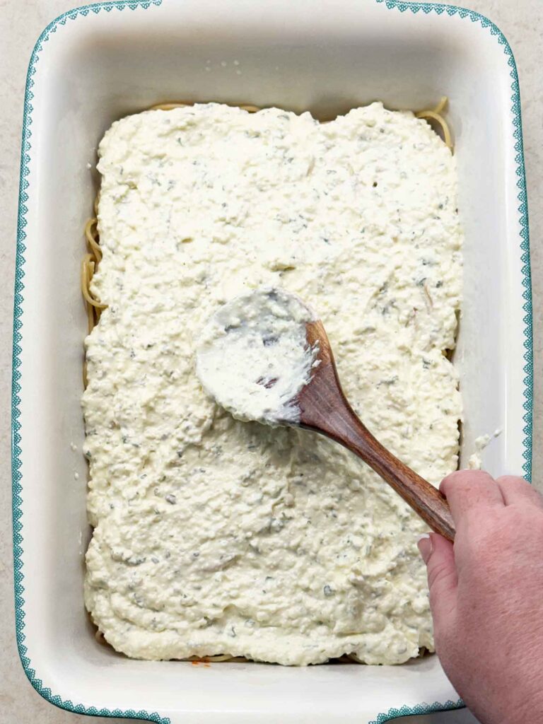 The creamy cottage cheese and cream cheese mixture being spread over the spaghetti with a wooden spoon.
