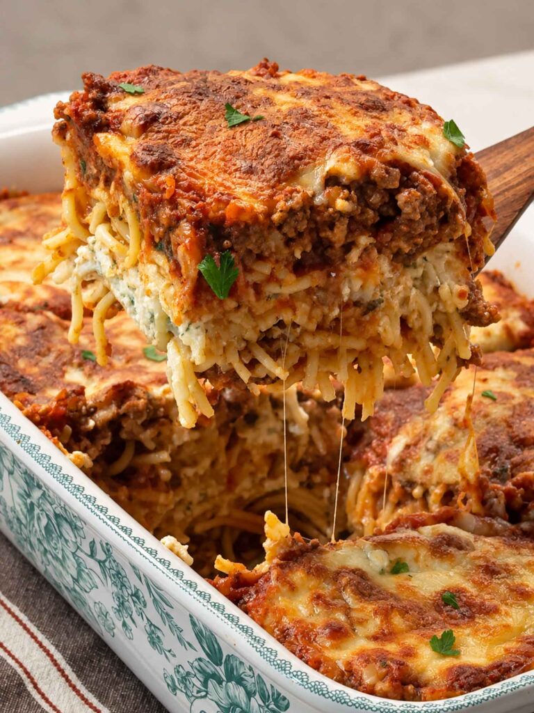 A square of million dollar spaghetti lifted out of the baking dish on a wooden spoon, cheese stretching, showing layers of spaghetti, cottage cheese filling, and meat sauce.