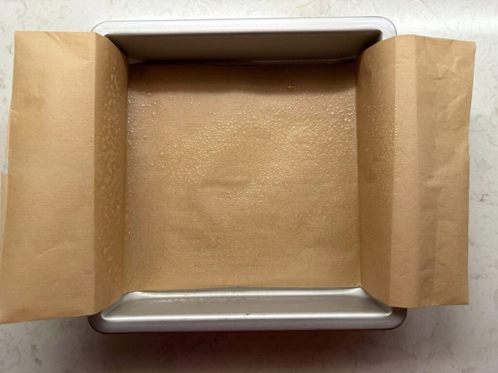 An 8x8 metal baking pan lined with a strip of parchment paper that hangs over two sides