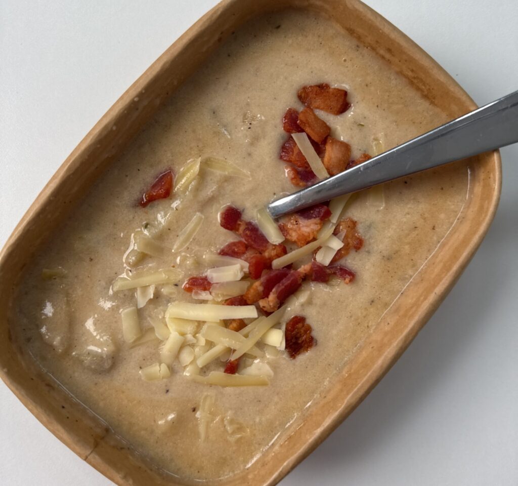 A paper meal prep container full of potato soup with bacon and cheese sprinkled on top