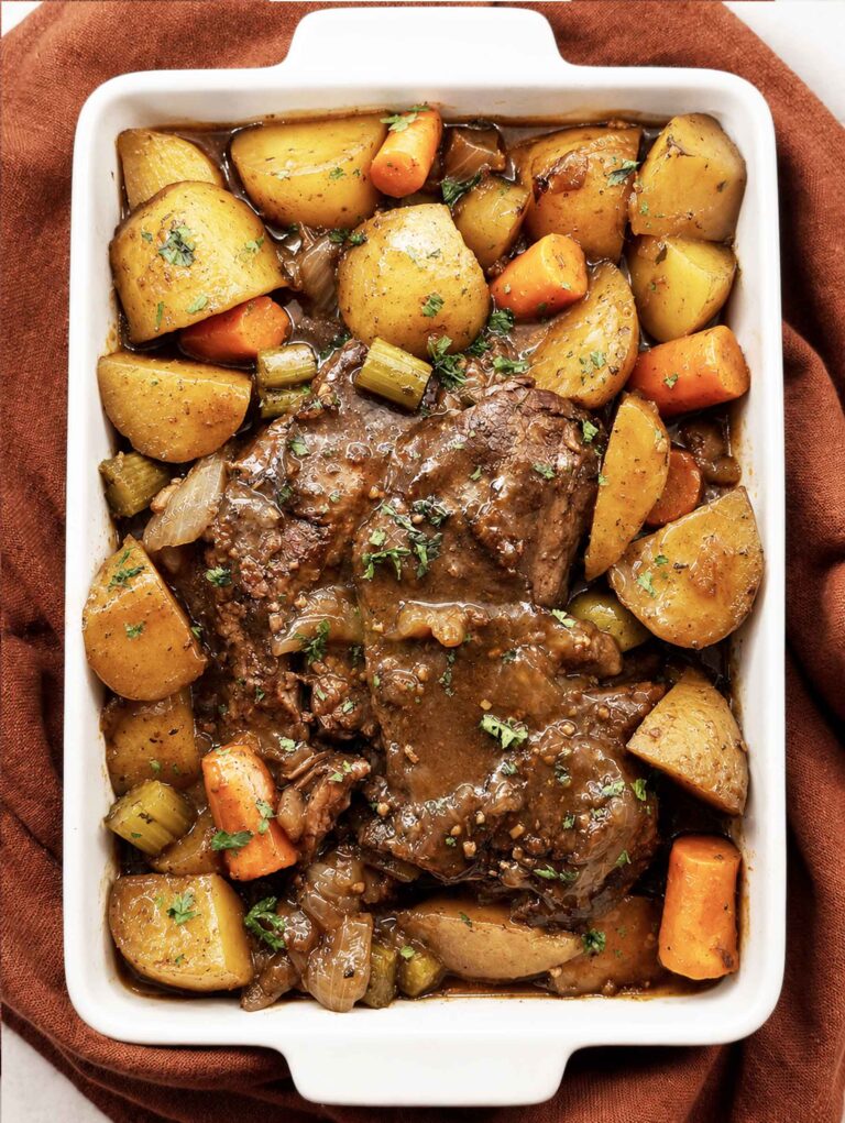 Overhead view of slow cooker pot roast in a white serving dish with potatoes, carrots, and celery in rich brown gravy