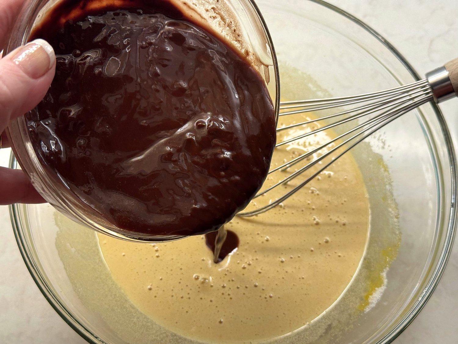 Melted chocolate being poured from a glass bowl into the whisked eggs and sugar