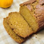 Sliced loaf of homemade pumpkin bread on a beige gingham cloth with a small orange pumpkin