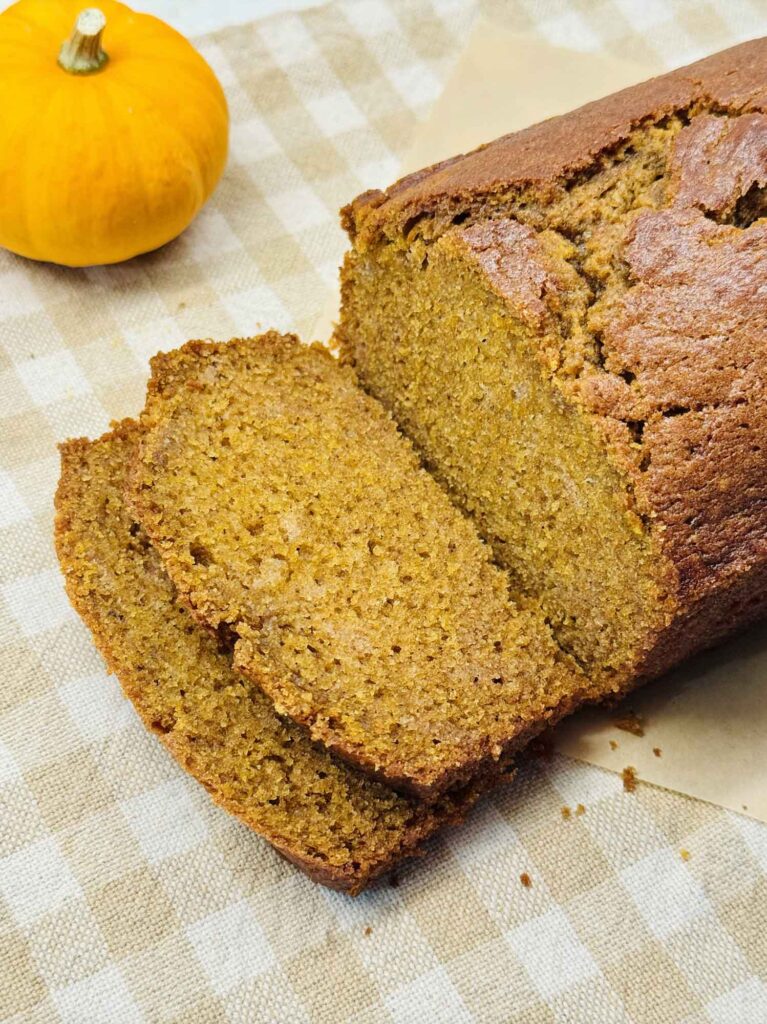 Sliced loaf of homemade pumpkin bread on a beige gingham cloth with a small orange pumpkin