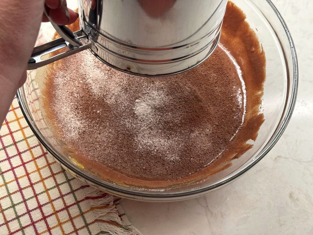 A hand-crank flour sifter sifting flour, cocoa powder, and salt over chocolate batter in a glass bowl