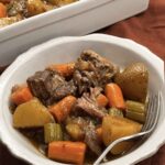 A served bowl of slow cooker pot roast with fork-tender beef, potatoes, carrots, and celery in gravy, serving dish behind