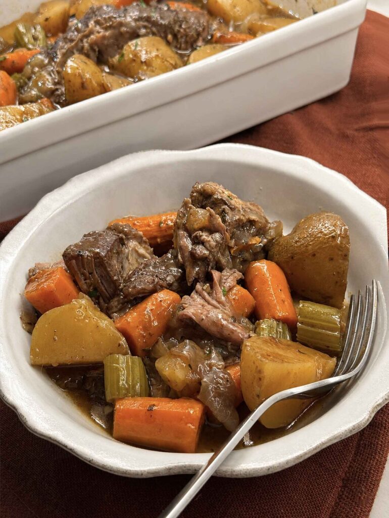 A served bowl of slow cooker pot roast with fork-tender beef, potatoes, carrots, and celery in gravy, serving dish behind