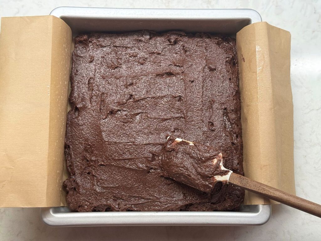 Thick brownie batter being spread into the corners of a parchment-lined 8x8 pan with a spatula