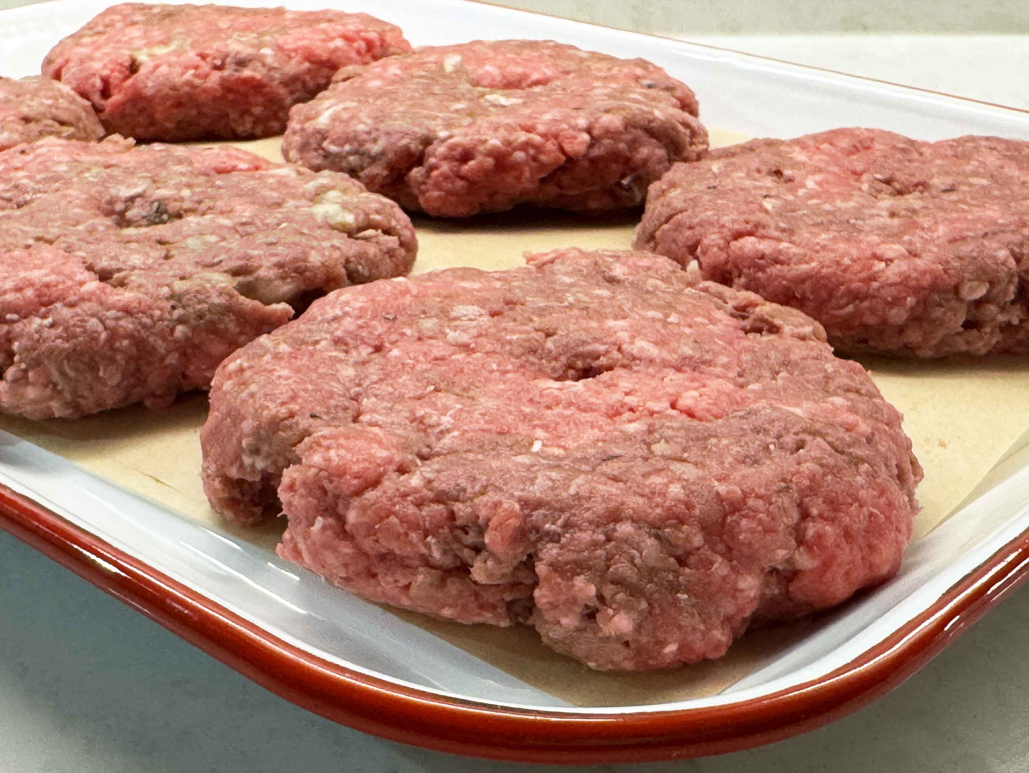 Six raw burger patties with thumb dimples resting on parchment paper