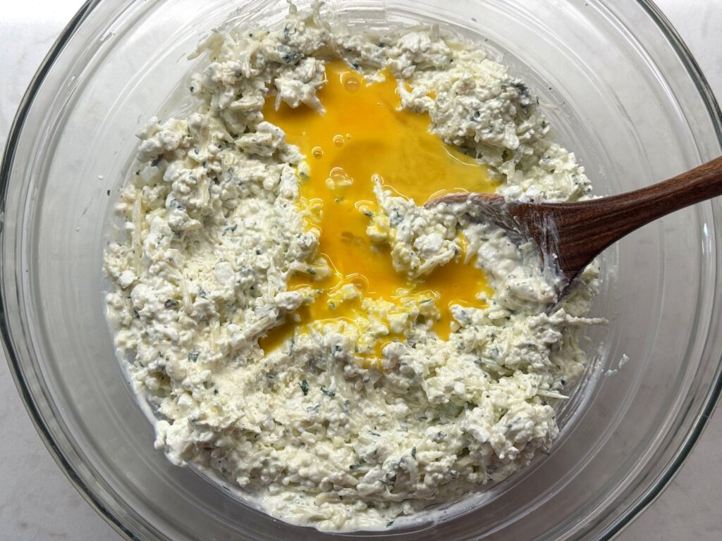 A beaten egg on top of mixed cheese filling in a glass bowl with a wooden spoon