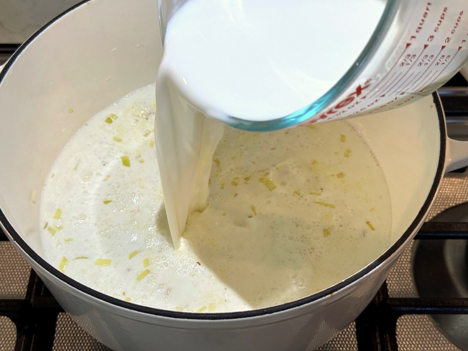 Half-and-half being poured from a glass measuring cup over softened onions in a Dutch oven