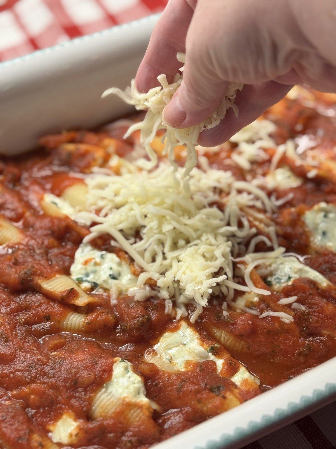 A hand sprinkling shredded mozzarella over saucy stuffed shells