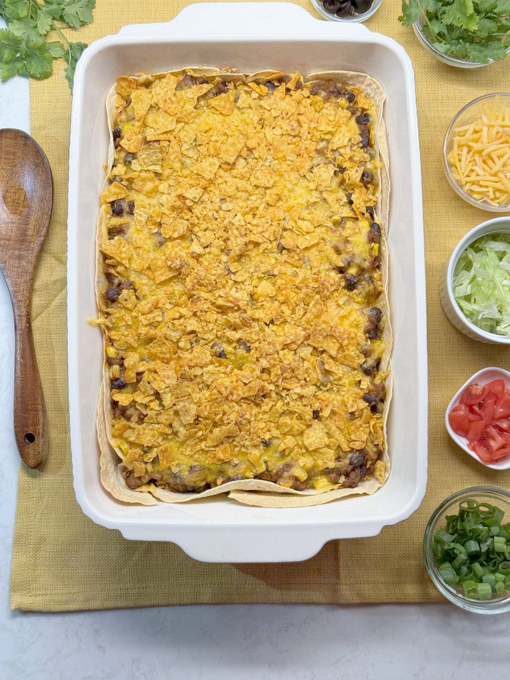 Finished taco casserole with a toasted Doritos top in a white baking dish, surrounded by bowls of toppings