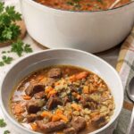 A gray bowl of beef barley soup with tender beef, diced carrots, and barley, in front of a white Dutch oven