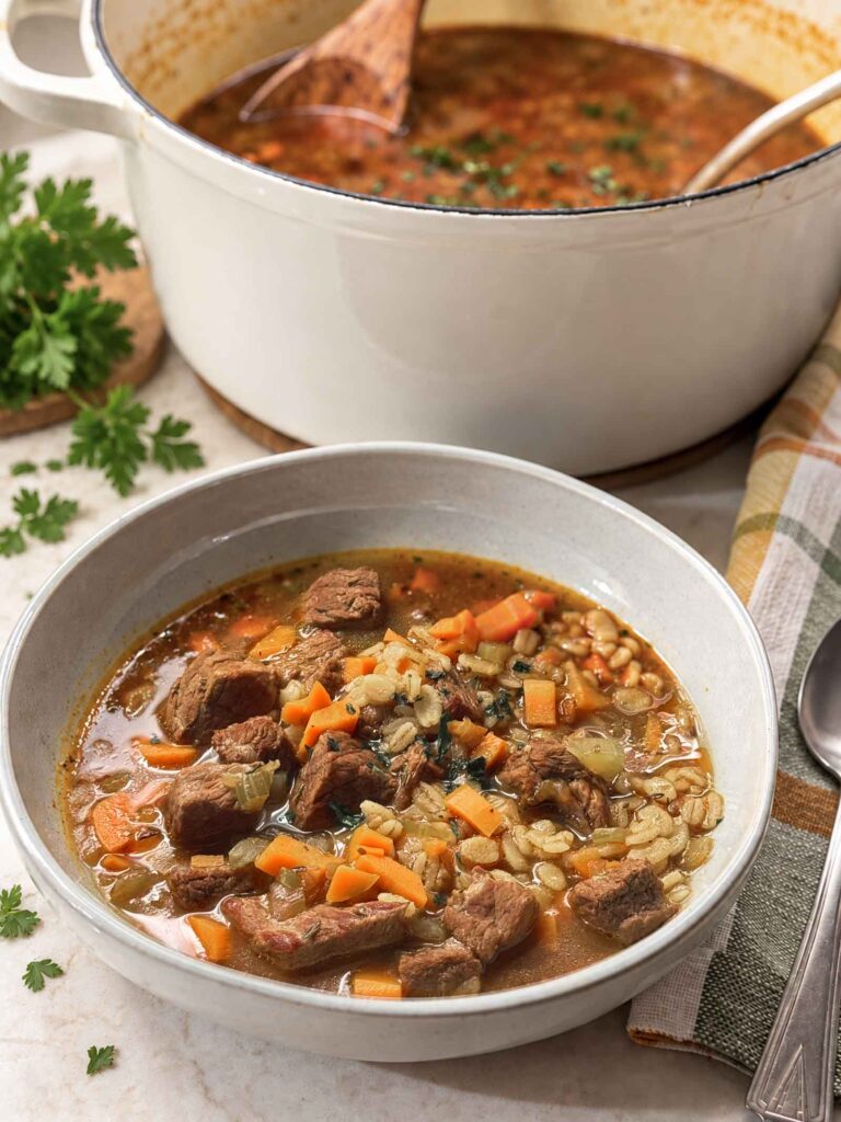 A gray bowl of beef barley soup with tender beef, diced carrots, and barley, in front of a white Dutch oven