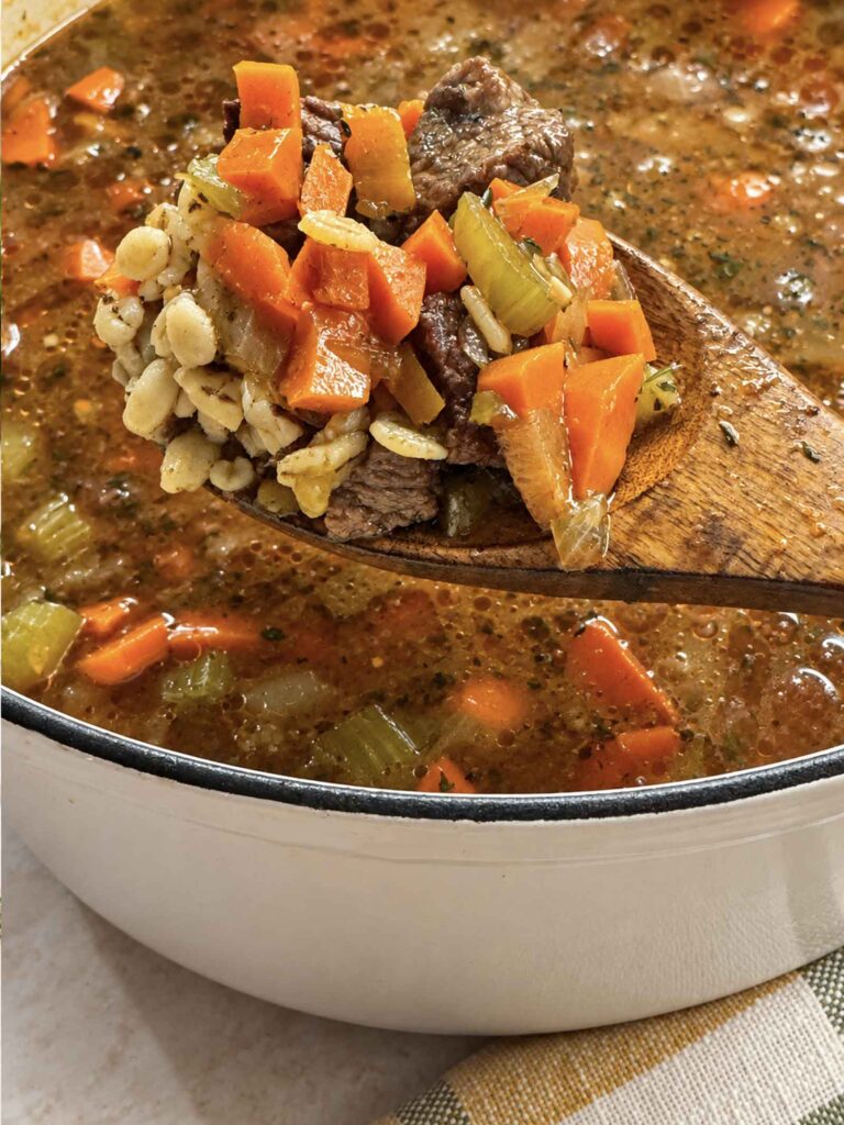 A wooden spoon lifting beef, carrots, celery, and cooked barley out of the pot