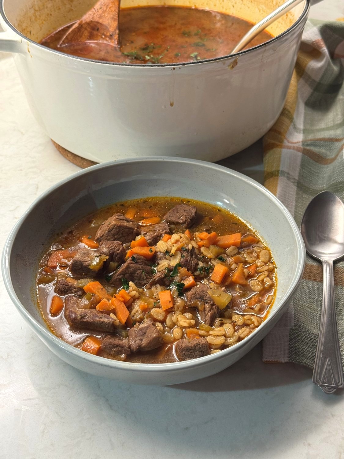 A bowl of beef barley soup with a spoon, a plaid towel, and the Dutch oven behind it