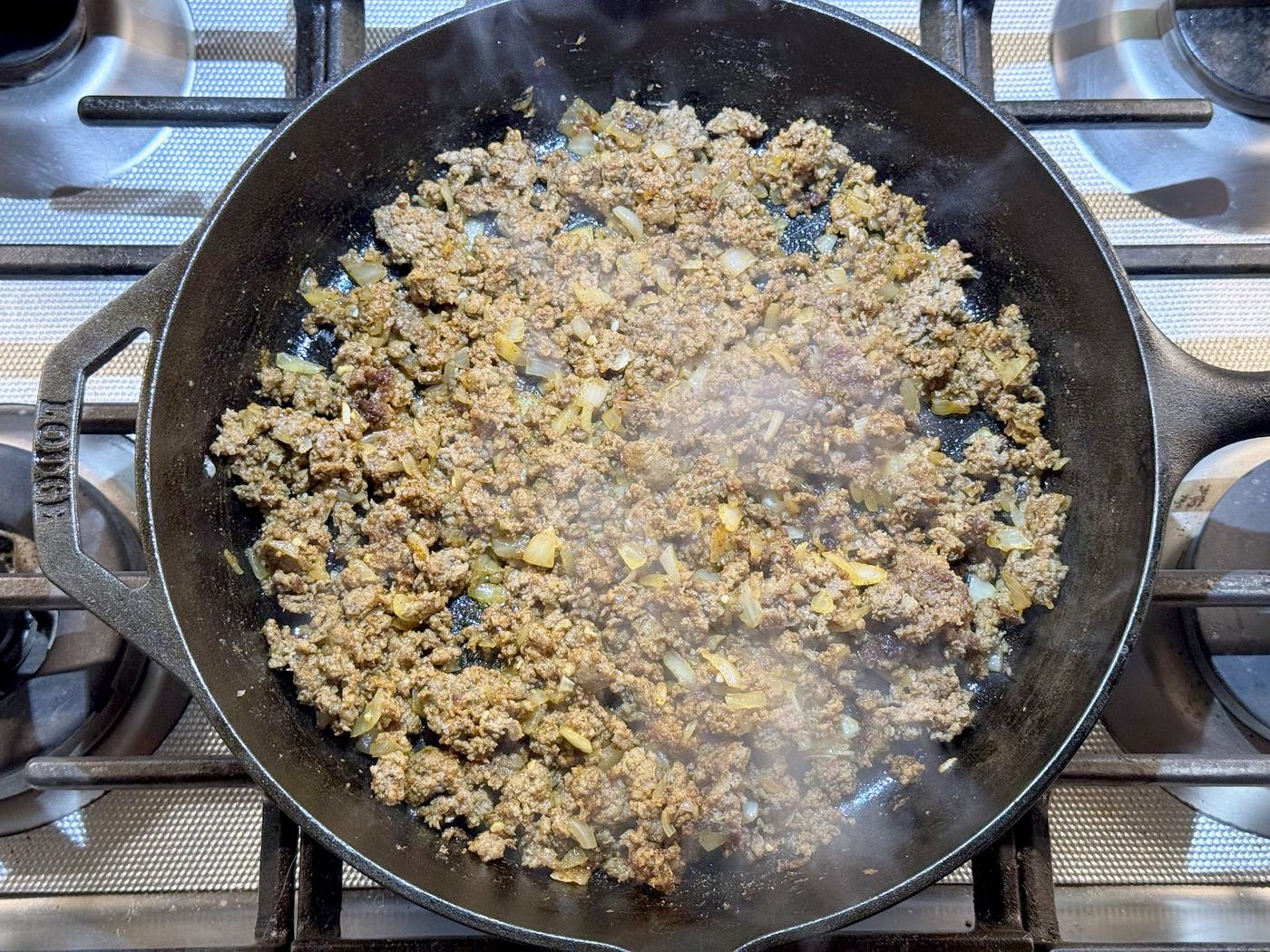 Ground beef and diced onion browning in a cast iron skillet on a gas stove