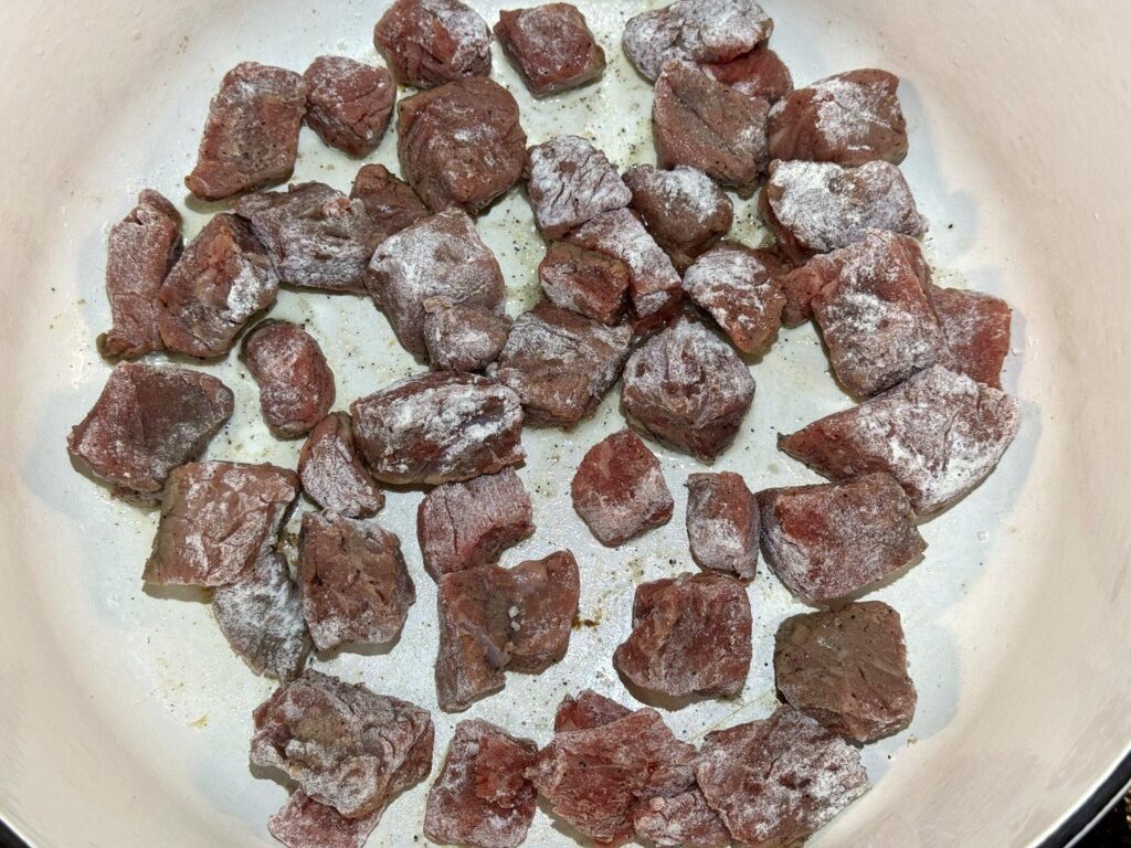 Flour-coated stew meat in a single layer in a white Dutch oven
