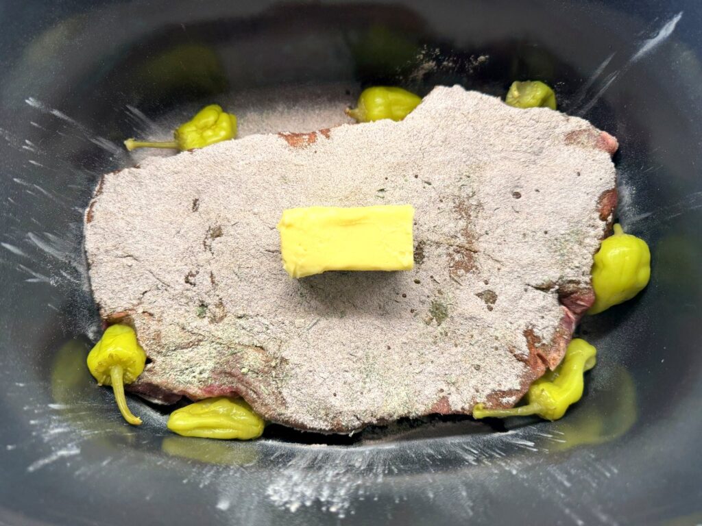 Half a stick of butter on a seasoned chuck roast with whole pepperoncini tucked around it in a dry slow cooker