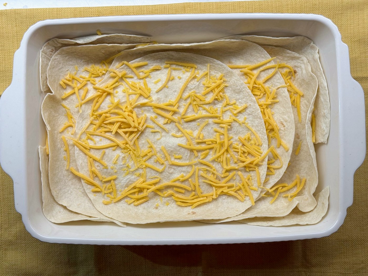 Shredded cheddar sprinkled over flour tortillas lining a white baking dish