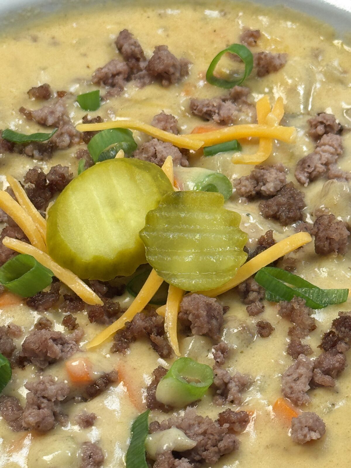 Close-up of dill pickle slices, shredded cheddar, green onions, and ground beef crumbles on top of creamy cheeseburger soup
