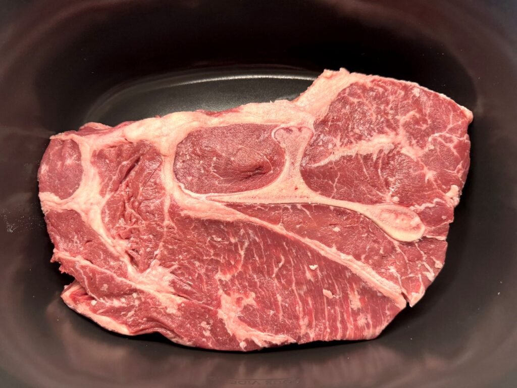A raw bone-in beef chuck roast lying flat in an oval slow cooker