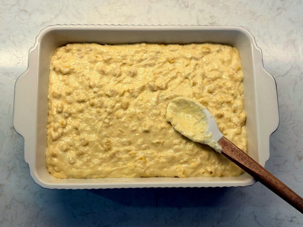 Corn casserole batter spread evenly in a white 7x10 baking dish with a spatula