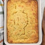 Overhead view of golden baked corn casserole in a white baking dish on an orange plaid towel with a wooden spatula