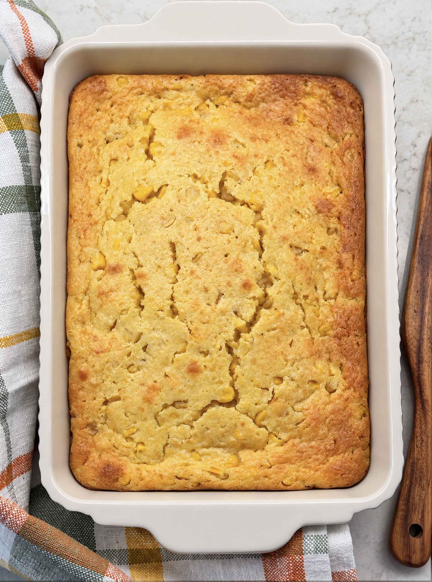 Overhead view of golden baked corn casserole in a white baking dish on an orange plaid towel with a wooden spatula