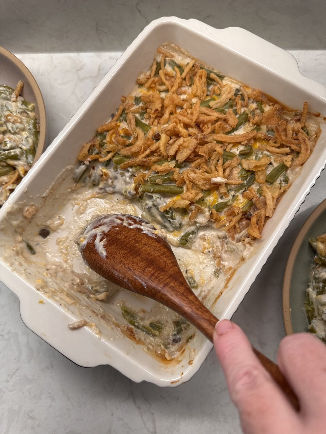 A wooden spoon scooping creamy green bean casserole from the corner of the baking dish