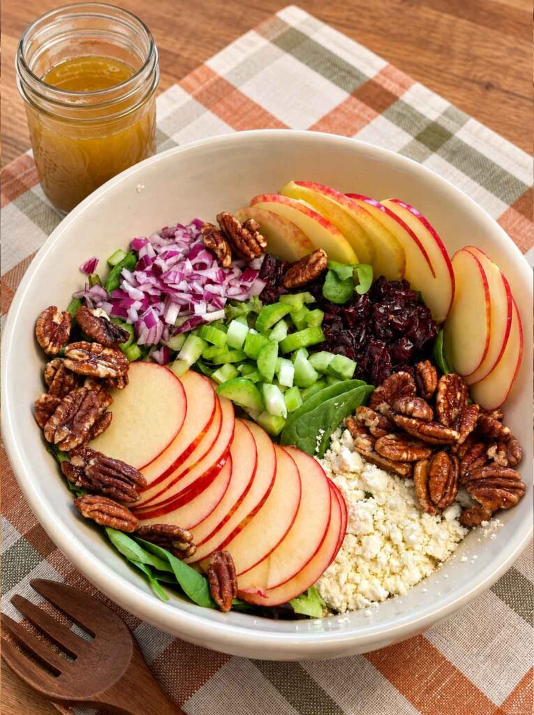 A bowl of salad with apples, feta cheese, maple pecans, red onions, celery and greens, and a mason jar of maple vinaigrette nearby