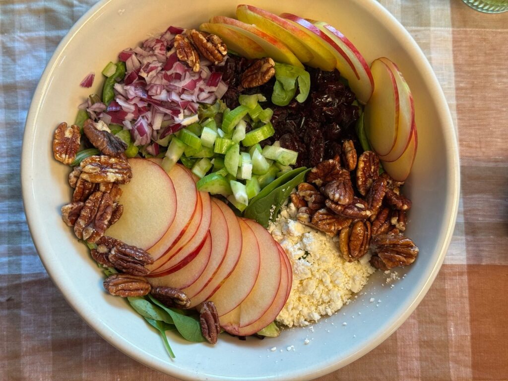Overhead view of a fall salad in a white bowl with apple slices, maple pecans, feta, cranberries, celery, and red onion arranged in sections over spinach