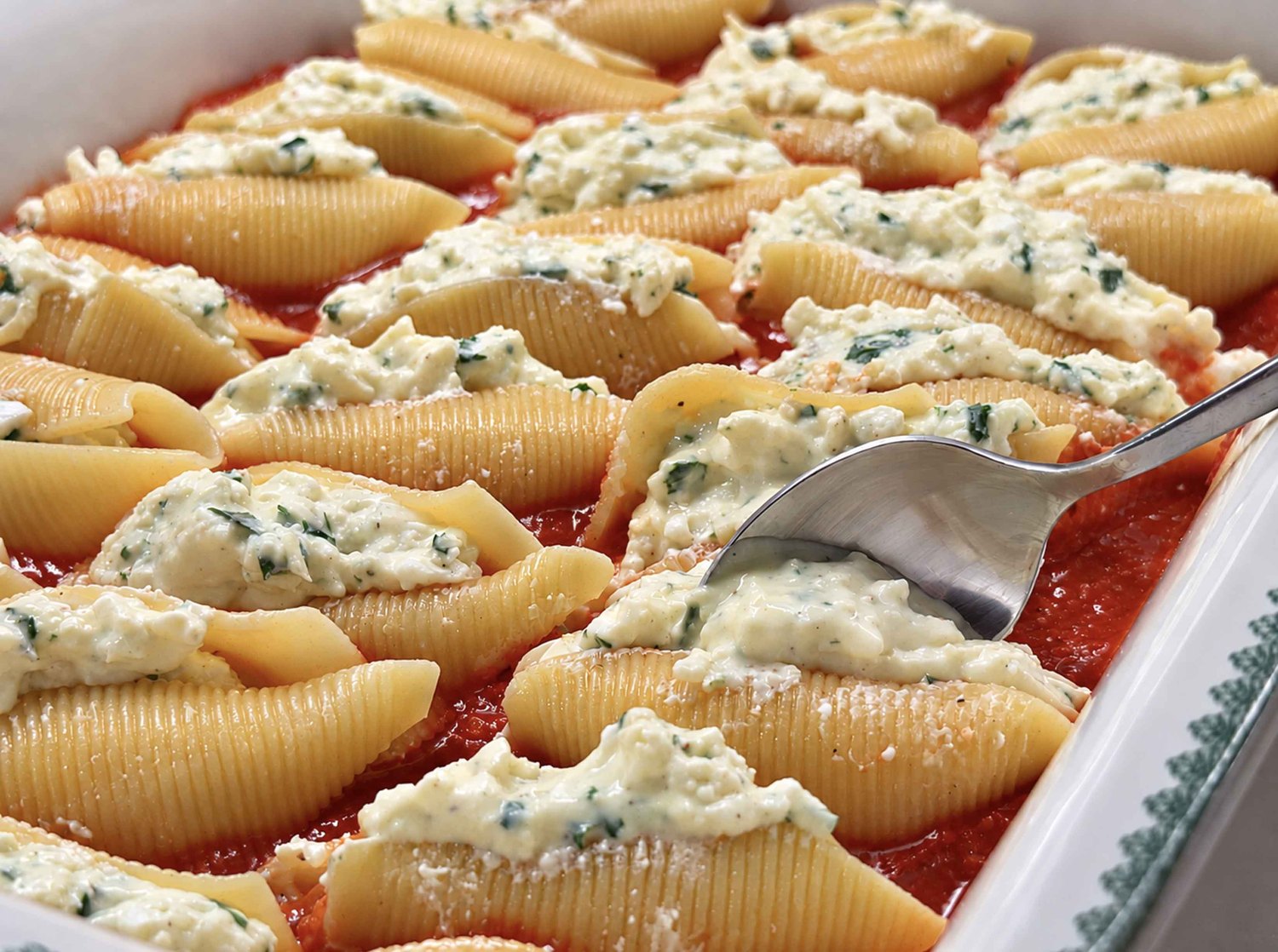 Spooning creamy cheese filling into jumbo shells in a baking dish of marinara