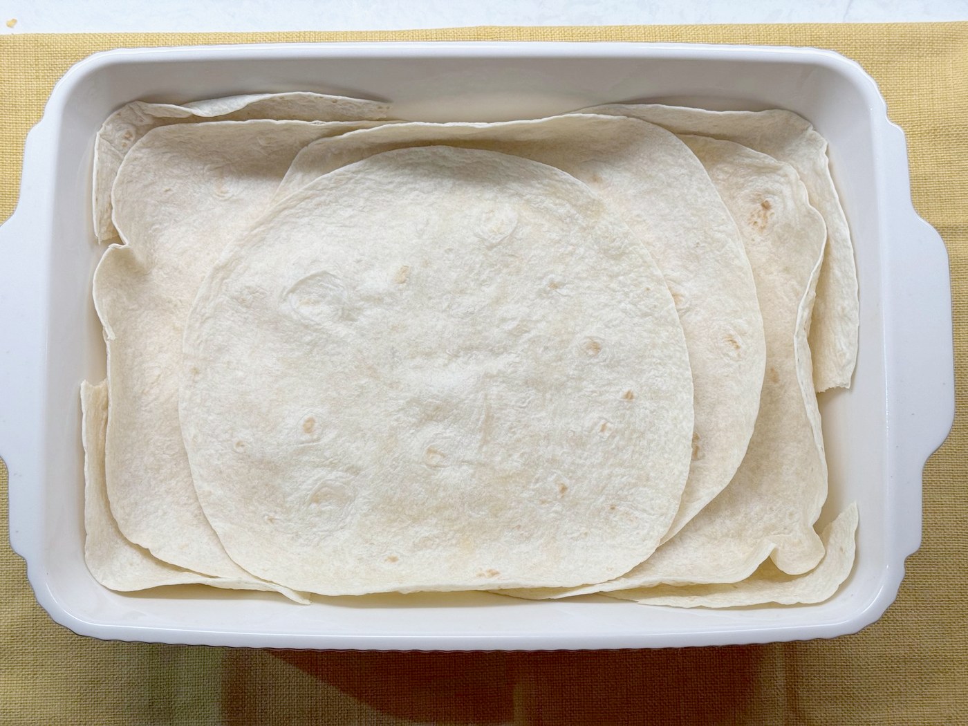 Five flour tortillas overlapping to cover the bottom of a white 9x13 baking dish, with the edges coming up the sides