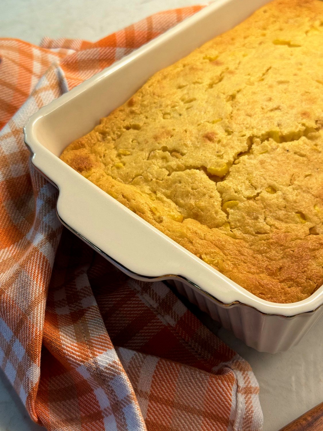 Baked corn casserole in a white baking dish on an orange plaid towel, seen at an angle
