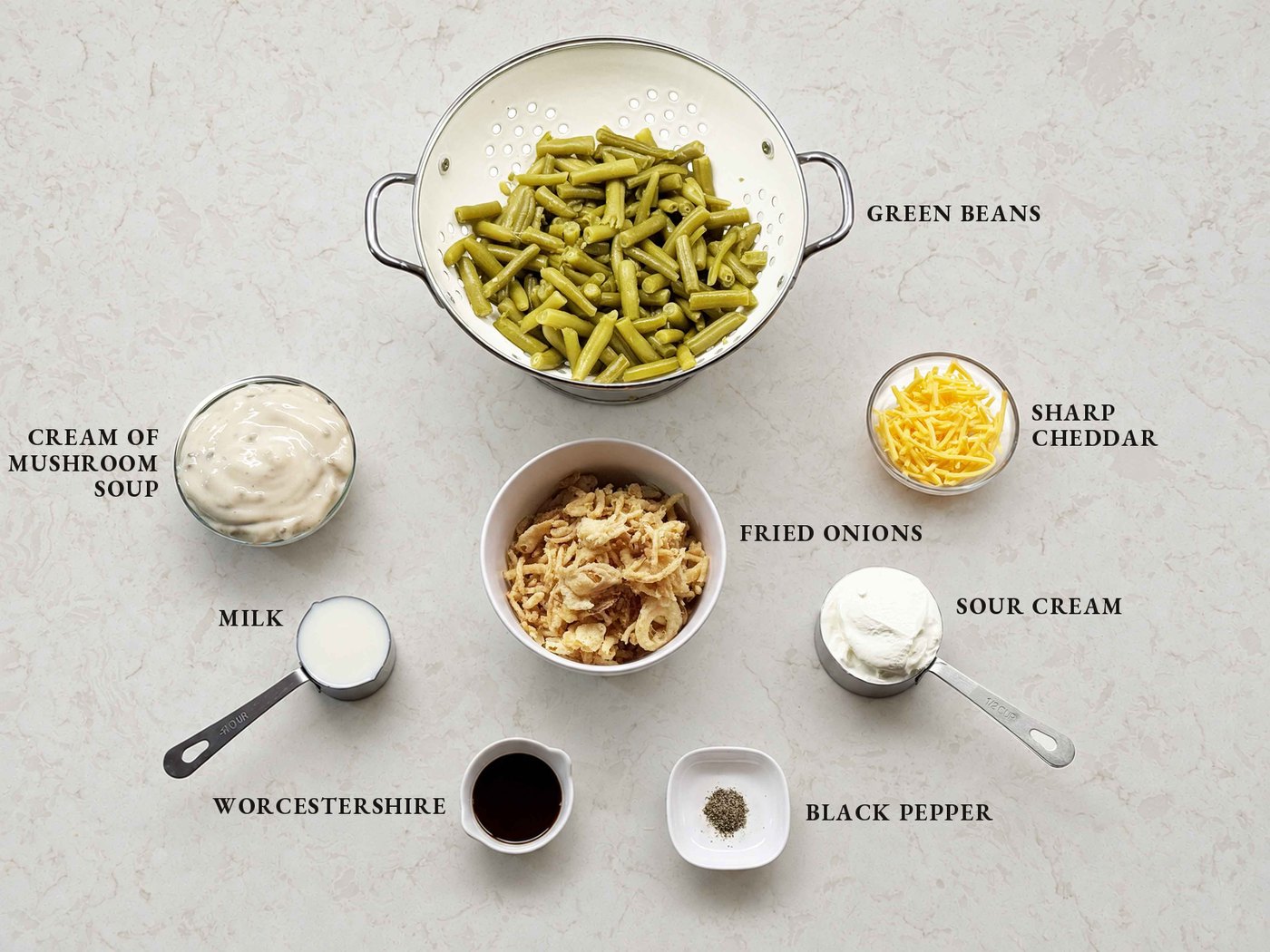 Labeled ingredients for green bean casserole: drained green beans, cream of mushroom soup, sharp cheddar, fried onions, sour cream, milk, Worcestershire sauce, and black pepper